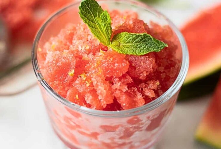 A beautifully plated watermelon lime granita served in a chilled glass bowl, garnished with a fresh lime wheel and mint sprig, vibrant red-pink crystals glistening in natural sunlight on a white marble surface