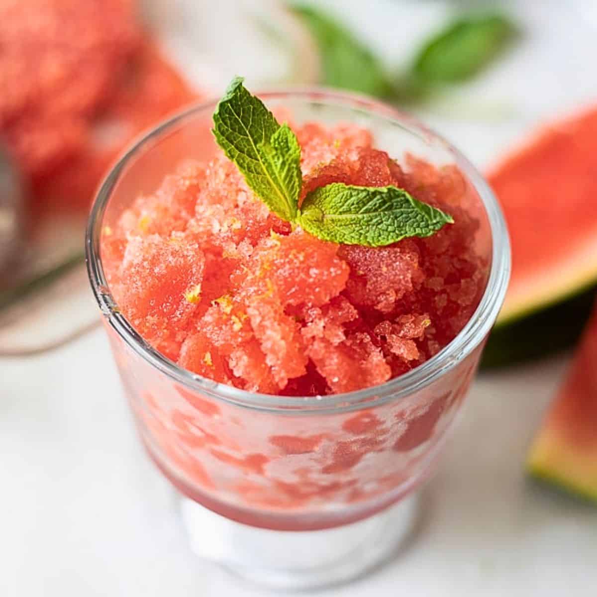 A beautifully plated watermelon lime granita served in a chilled glass bowl, garnished with a fresh lime wheel and mint sprig, vibrant red-pink crystals glistening in natural sunlight on a white marble surface
