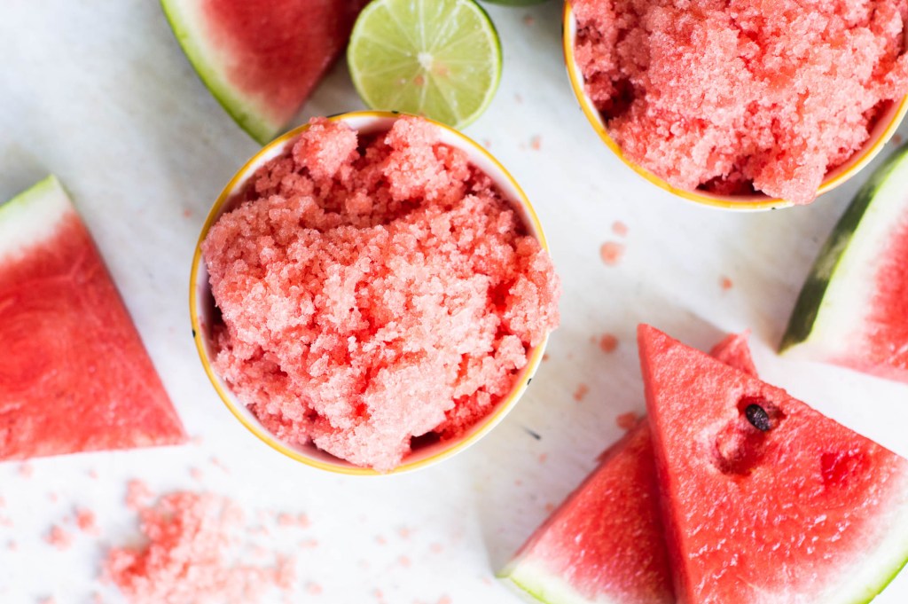 Overhead flat lay of watermelon lime granita ingredients — cubed fresh watermelon, halved limes, raw honey in a small jar, fresh mint leaves, and sea salt on a light linen surface, with a blender and shallow baking dish nearby