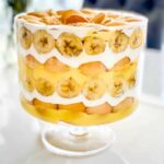 Classic banana pudding served in a wide glass trifle bowl, showing beautiful layers of vanilla wafers, fresh banana slices, and creamy custard topped with billowy whipped cream and wafer crumbles