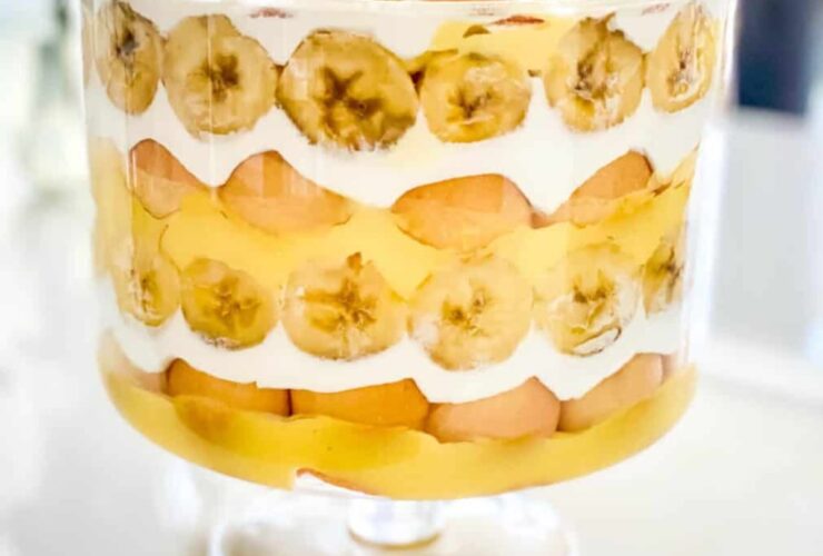 Classic banana pudding served in a wide glass trifle bowl, showing beautiful layers of vanilla wafers, fresh banana slices, and creamy custard topped with billowy whipped cream and wafer crumbles