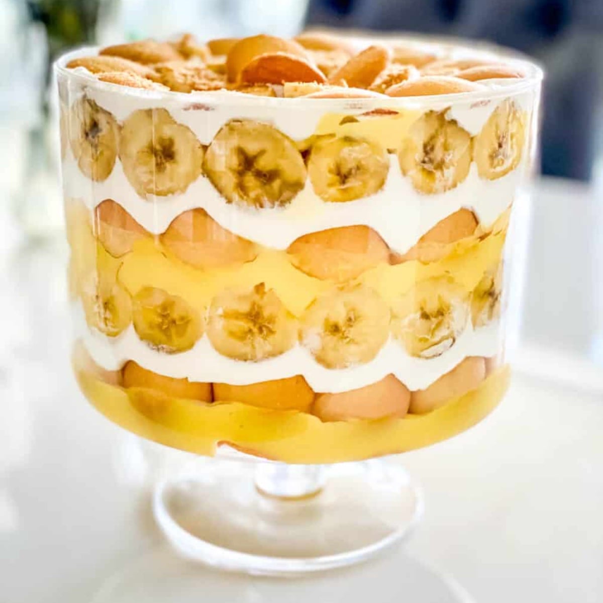 Classic banana pudding served in a wide glass trifle bowl, showing beautiful layers of vanilla wafers, fresh banana slices, and creamy custard topped with billowy whipped cream and wafer crumbles