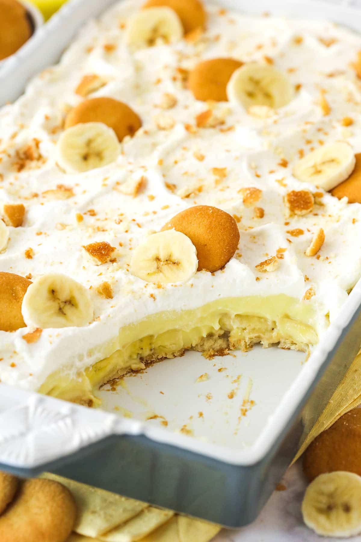 Step-by-step ingredients flat lay showing ripe spotted bananas, vanilla wafers in a box, eggs, whole milk, vanilla extract, sugar, and a bowl of freshly whipped cream on a white marble surface