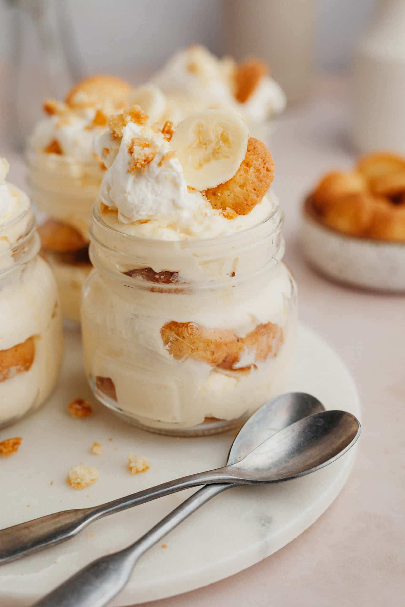 Close-up overhead shot of a single serving of banana pudding in a mason jar, showing distinct layers of golden custard, white vanilla wafers, fresh banana slices, and a swirl of whipped cream topped with a whole vanilla wafer and banana chip garnish