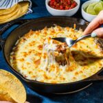 A rustic cast iron skillet filled with creamy seasoned beans topped with golden melted cheese, garnished with fresh cilantro, diced avocado, and a squeeze of lime on a warm wooden table