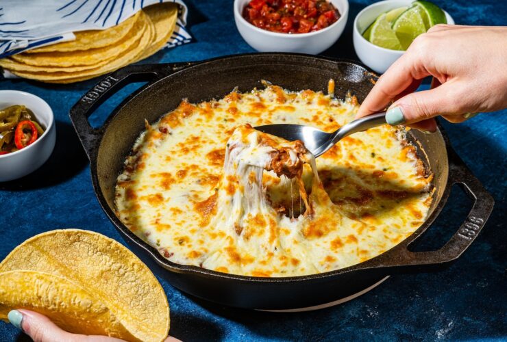 A rustic cast iron skillet filled with creamy seasoned beans topped with golden melted cheese, garnished with fresh cilantro, diced avocado, and a squeeze of lime on a warm wooden table