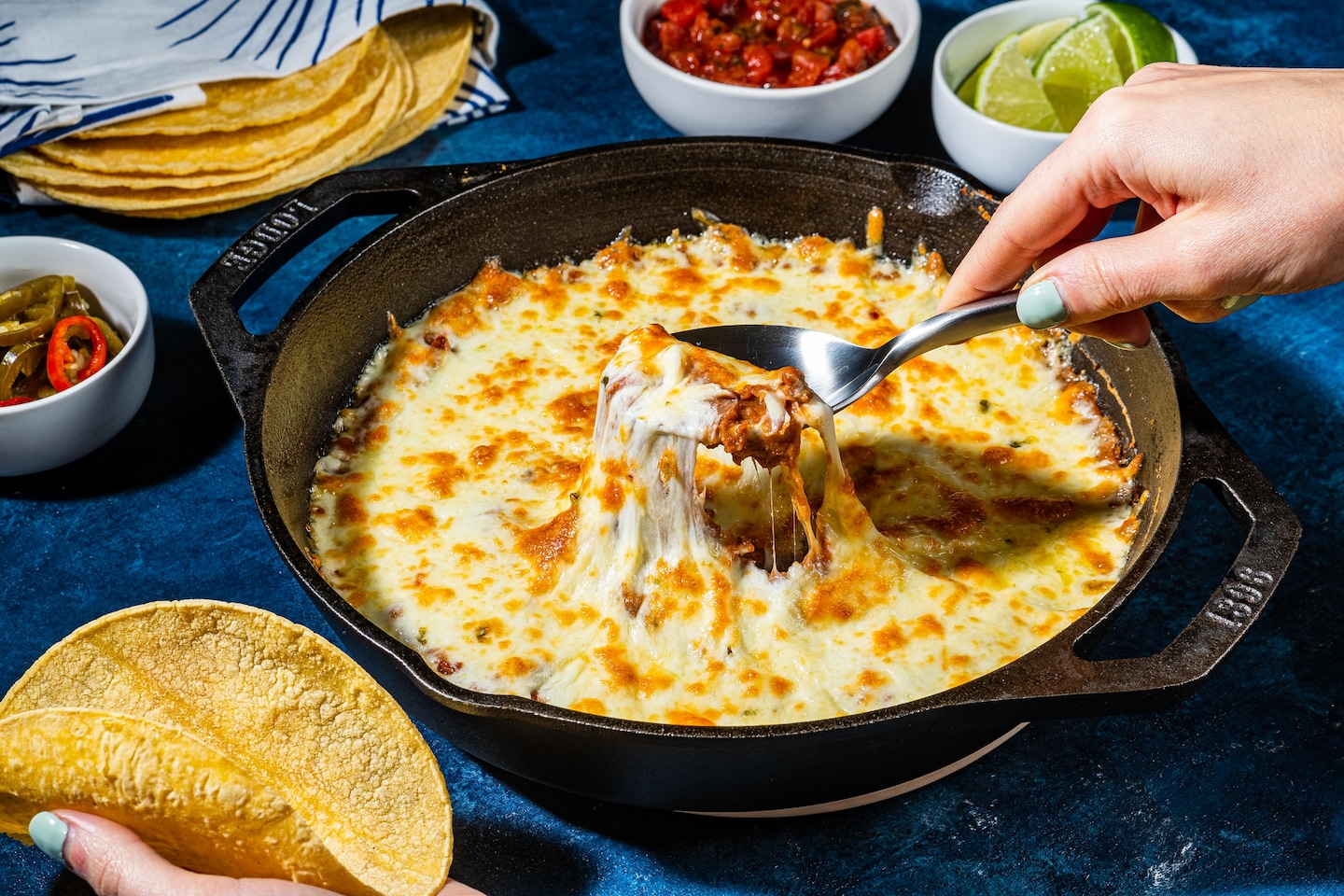 A rustic cast iron skillet filled with creamy seasoned beans topped with golden melted cheese, garnished with fresh cilantro, diced avocado, and a squeeze of lime on a warm wooden table
