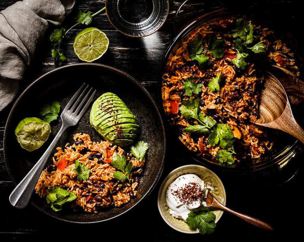 Overhead flat lay of key ingredients — two cans of black beans, a block of sharp cheddar, fresh garlic bulbs, a yellow onion, canned diced tomatoes, cumin, and smoked paprika arranged on a rustic cream linen surface