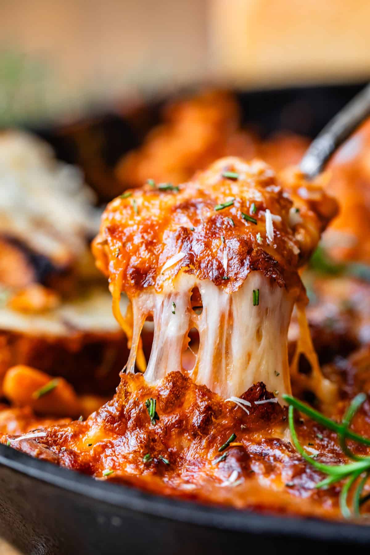 A close-up of a wooden spoon lifting a generous scoop of cheesy beans from a cast iron skillet, with melted cheddar stretching and fresh cilantro and lime wedges visible in the background