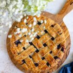 golden lattice top blueberry pie cooling on a wooden table surrounded by fresh blueberries and a vintage pie server