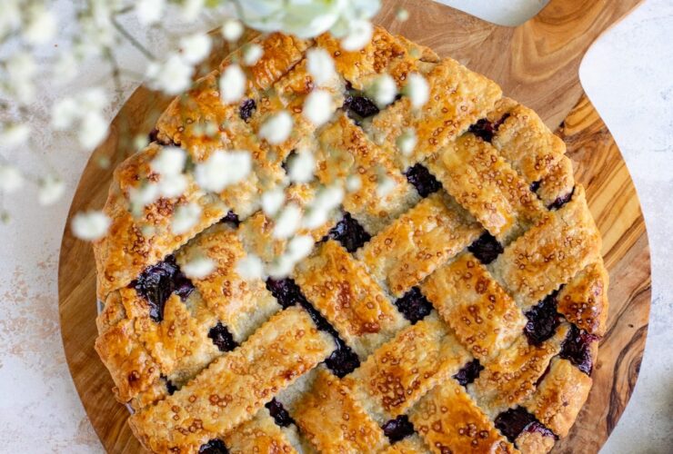 golden lattice top blueberry pie cooling on a wooden table surrounded by fresh blueberries and a vintage pie server