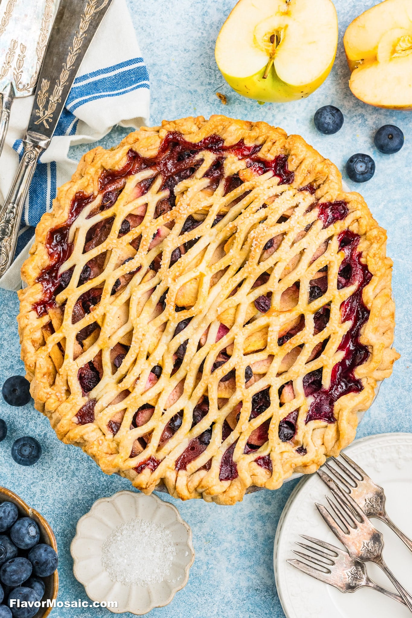 close-up of hands weaving a pie lattice crust over a bowl of fresh blueberries and measured ingredients on a marble surface