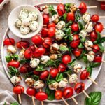 Caprese skewers beautifully plated on a white marble board with cherry tomatoes, fresh mozzarella, and basil leaves drizzled with balsamic glaze and olive oil