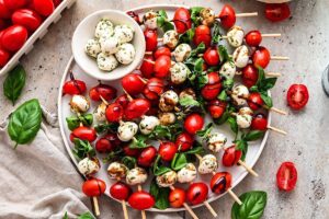 Caprese skewers beautifully plated on a white marble board with cherry tomatoes, fresh mozzarella, and basil leaves drizzled with balsamic glaze and olive oil