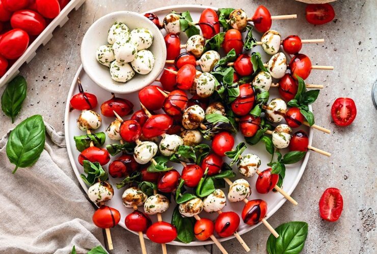 Caprese skewers beautifully plated on a white marble board with cherry tomatoes, fresh mozzarella, and basil leaves drizzled with balsamic glaze and olive oil
