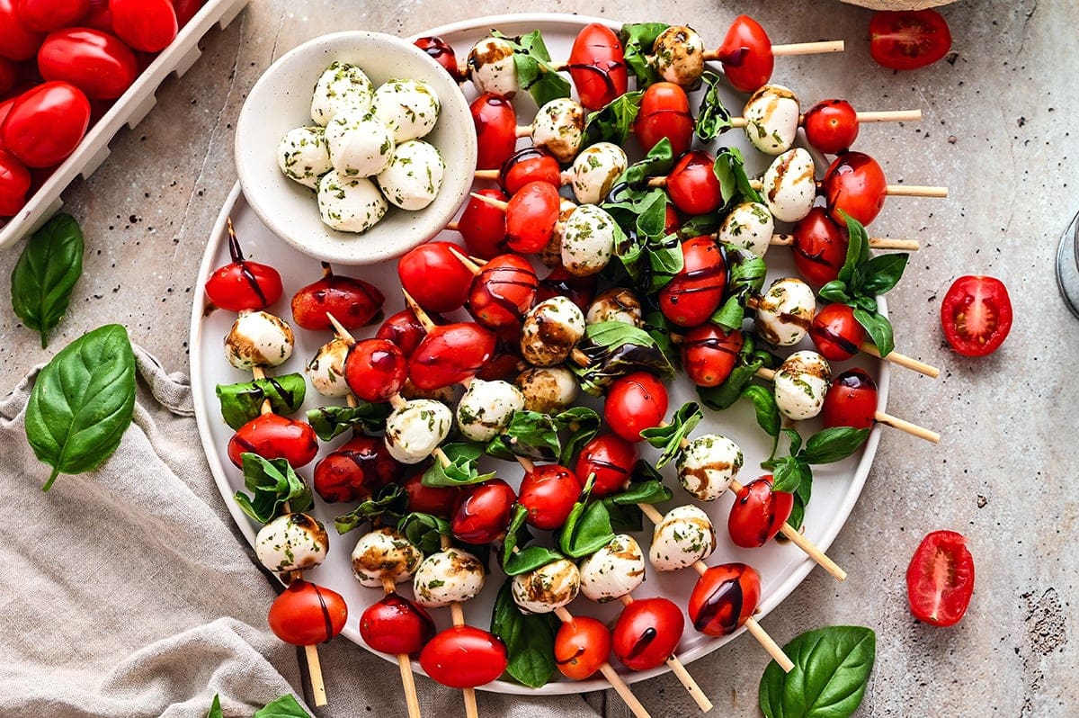 Caprese skewers beautifully plated on a white marble board with cherry tomatoes, fresh mozzarella, and basil leaves drizzled with balsamic glaze and olive oil
