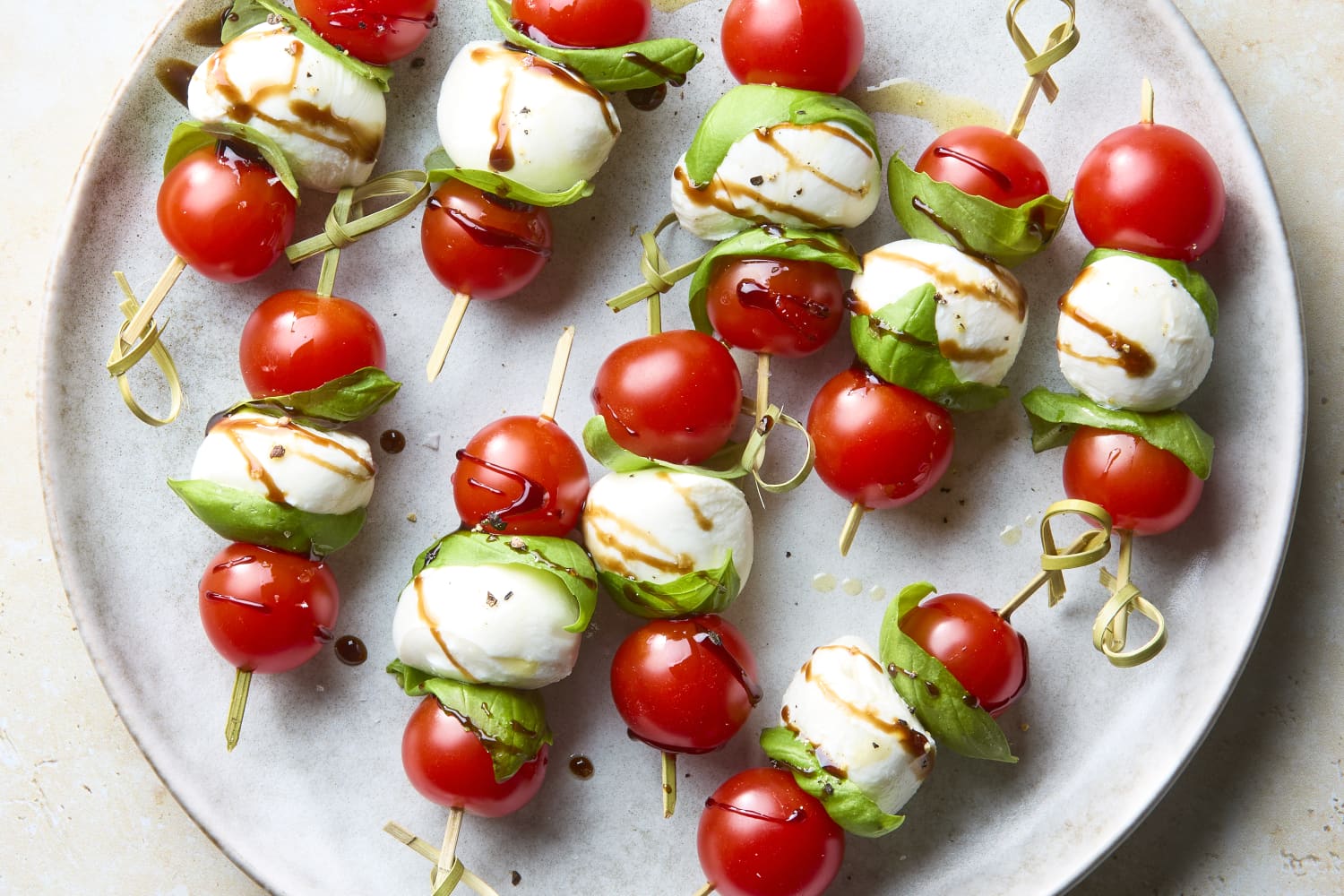 Caprese skewer ingredients laid out flat — cherry tomatoes, ciliegine mozzarella balls, fresh basil leaves, balsamic glaze, and olive oil on a light wood surface