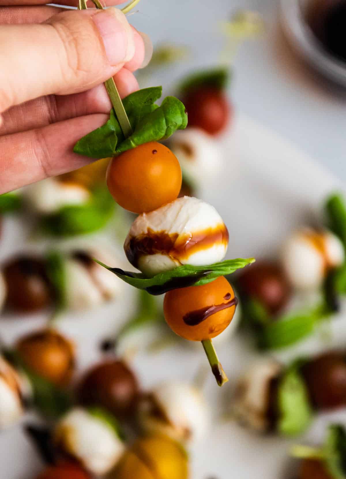 Close-up of a single Caprese skewer being held up with balsamic glaze dripping from the tomato, bright basil leaf and mozzarella visible below against a soft blurred background