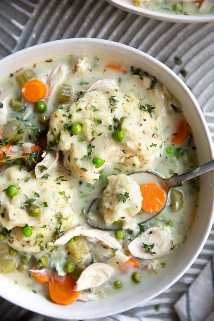 A close-up overhead shot of a rustic bowl of chicken and dumplings with a spoon resting inside, steam rising, surrounded by a linen napkin and fresh thyme sprigs
