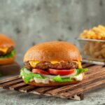 Classic Cheeseburgers stacked high with melted American cheese, crisp lettuce, tomato, and pickles on a toasted brioche bun, served on a wooden board at a sunny backyard cookout