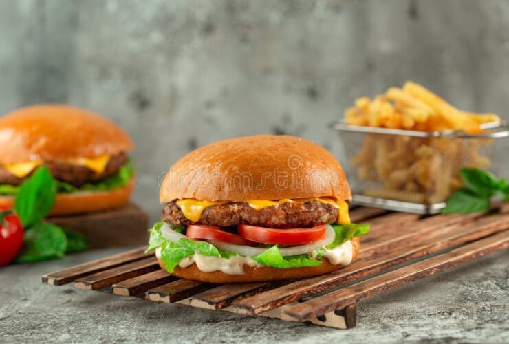 Classic Cheeseburgers stacked high with melted American cheese, crisp lettuce, tomato, and pickles on a toasted brioche bun, served on a wooden board at a sunny backyard cookout