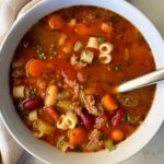 A steaming bowl of Crockpot Pasta e Fagioli with ground beef, cannellini beans, and ditalini pasta in a rich tomato broth, garnished with fresh parsley and Parmesan, photographed on a rustic wooden table