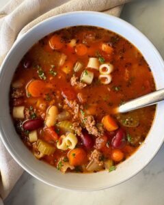 A steaming bowl of Crockpot Pasta e Fagioli with ground beef, cannellini beans, and ditalini pasta in a rich tomato broth, garnished with fresh parsley and Parmesan, photographed on a rustic wooden table