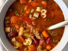A steaming bowl of Crockpot Pasta e Fagioli with ground beef, cannellini beans, and ditalini pasta in a rich tomato broth, garnished with fresh parsley and Parmesan, photographed on a rustic wooden table
