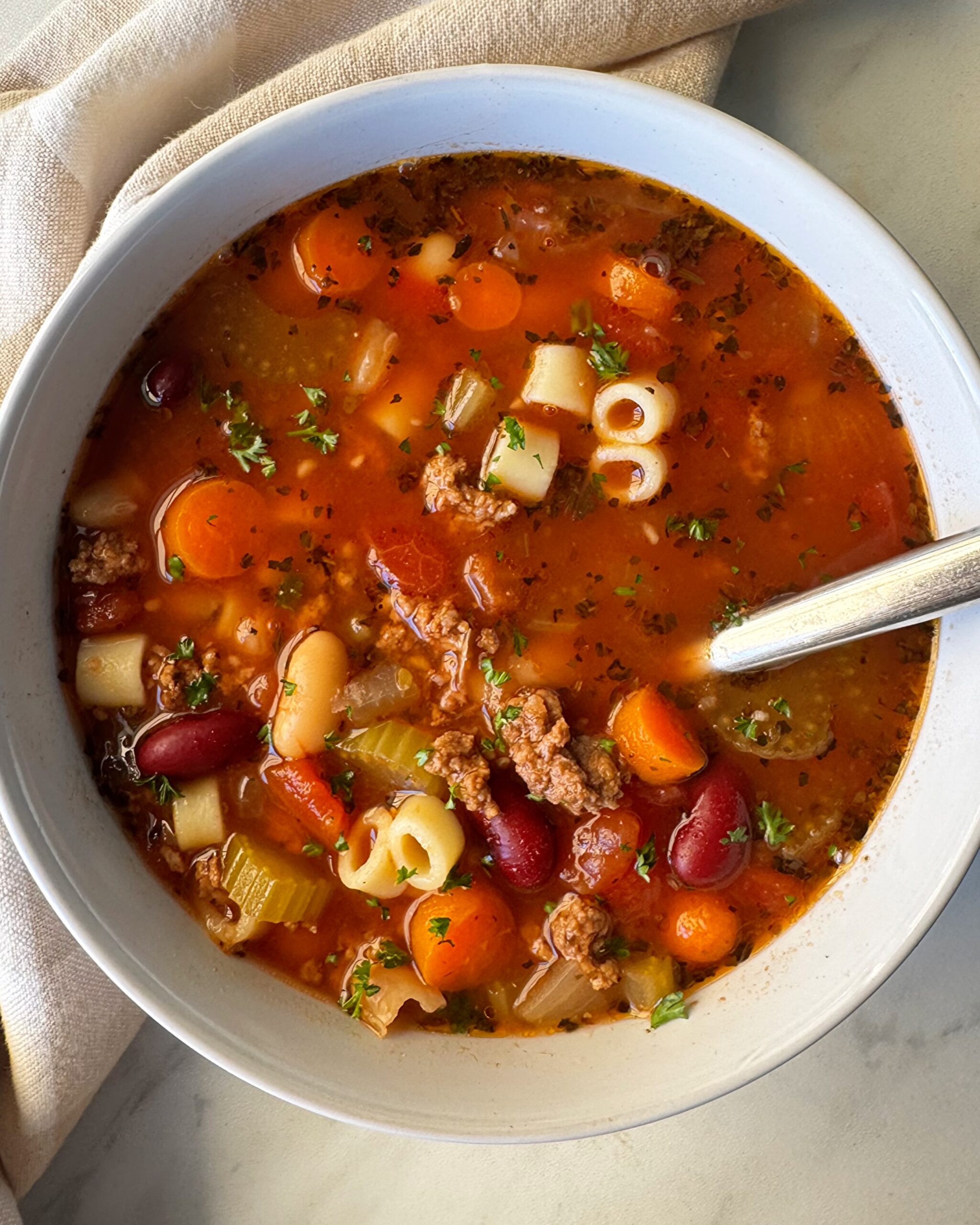 A steaming bowl of Crockpot Pasta e Fagioli with ground beef, cannellini beans, and ditalini pasta in a rich tomato broth, garnished with fresh parsley and Parmesan, photographed on a rustic wooden table