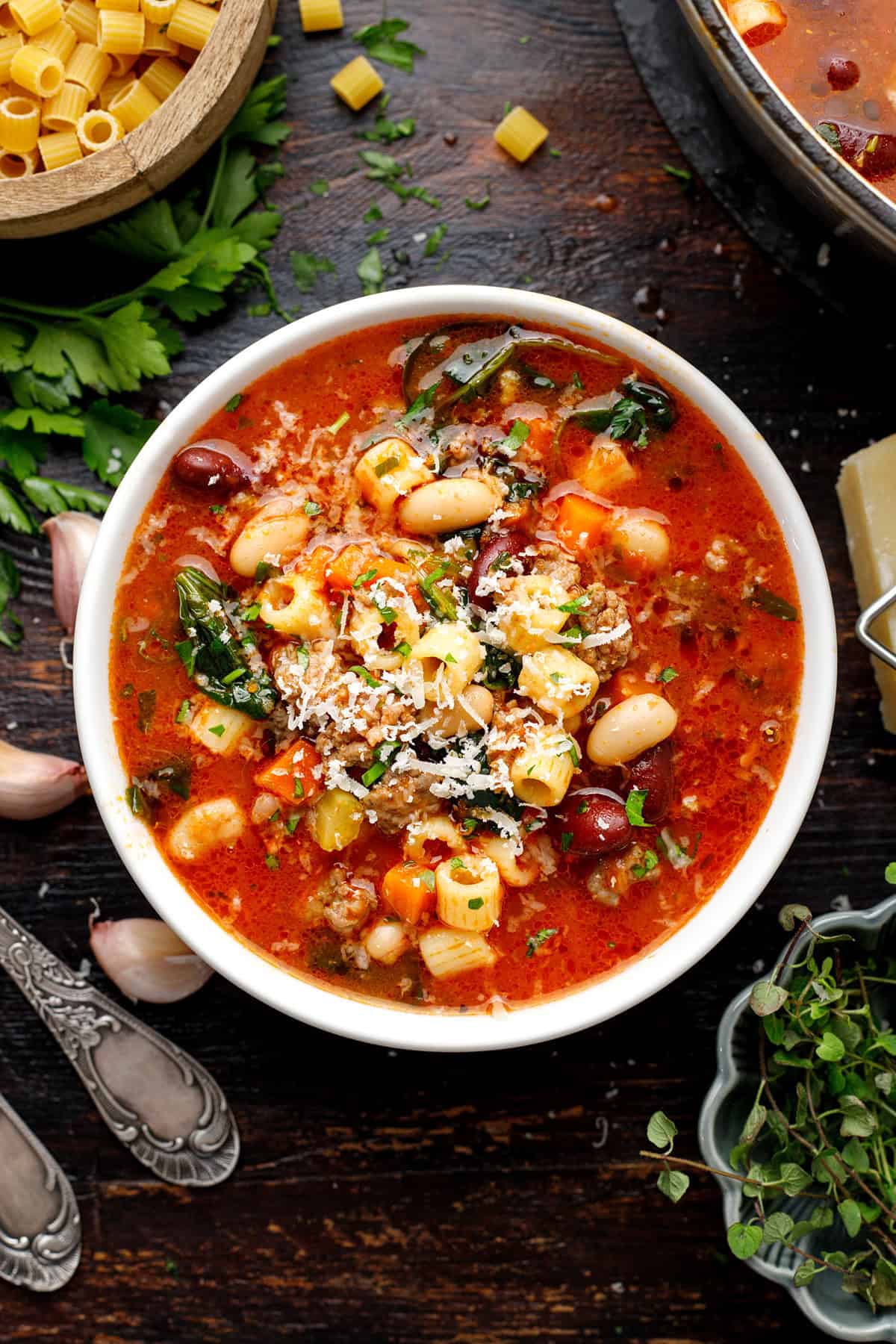 Overhead flat lay of Crockpot Pasta e Fagioli ingredients — cans of cannellini and kidney beans, crushed tomatoes, raw ground beef, ditalini pasta, celery, carrots, onion, garlic, and fresh herbs arranged on a white marble surface