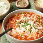 A rustic white bowl filled with Crockpot Stuffed Pepper Soup topped with shredded cheese and fresh parsley, set on a wooden table with a slow cooker in the background