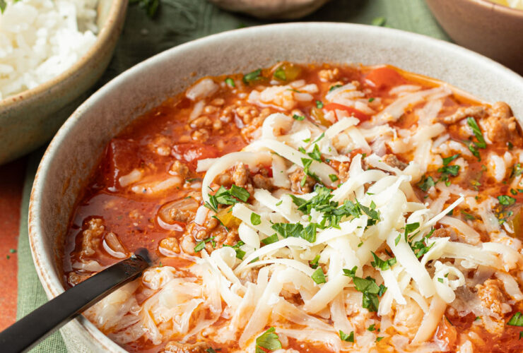 A rustic white bowl filled with Crockpot Stuffed Pepper Soup topped with shredded cheese and fresh parsley, set on a wooden table with a slow cooker in the background