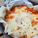 Dutch Oven Lasagna served from a cast iron Dutch oven at a campsite, bubbling with melted cheese and rich tomato sauce, golden and rustic
