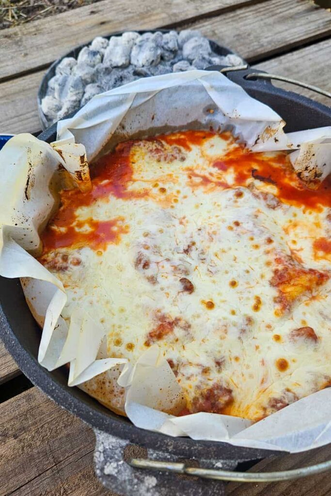 Dutch Oven Lasagna served from a cast iron Dutch oven at a campsite, bubbling with melted cheese and rich tomato sauce, golden and rustic