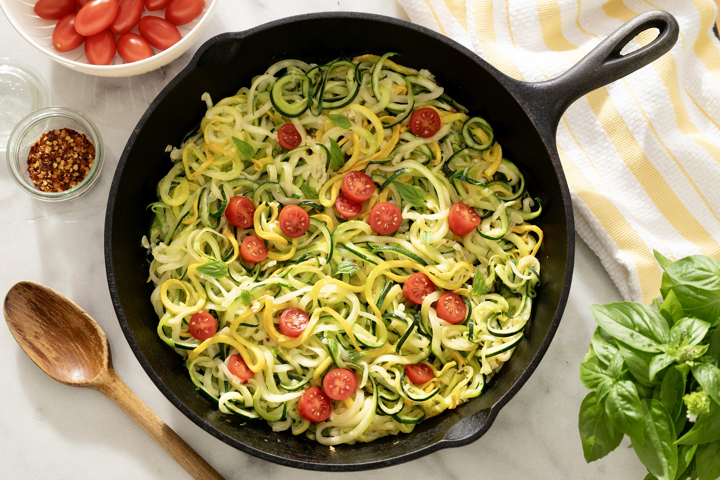 Overhead flat lay of spiralized zucchini noodles on a wooden cutting board beside fresh garlic cloves, a wedge of Parmesan, a stick of butter, red pepper flakes, and fresh parsley on a white marble surface