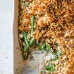 A beautifully plated green bean casserole in a white ceramic baking dish, topped with golden crispy fried onions, set on a rustic wooden table with autumn leaves in the background