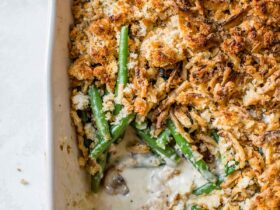 A beautifully plated green bean casserole in a white ceramic baking dish, topped with golden crispy fried onions, set on a rustic wooden table with autumn leaves in the background