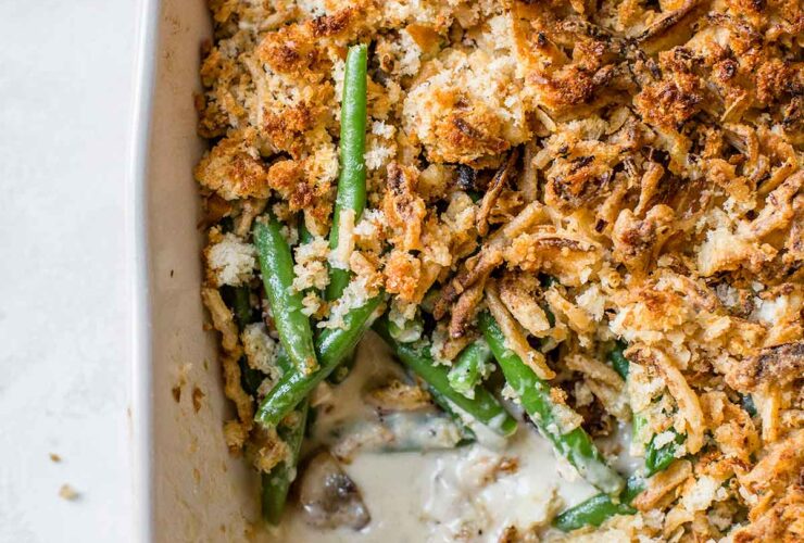A beautifully plated green bean casserole in a white ceramic baking dish, topped with golden crispy fried onions, set on a rustic wooden table with autumn leaves in the background