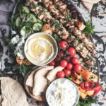 Grilled chicken skewers with tzatziki served on a wooden platter with pita, lemon wedges, and fresh herbs at a campsite