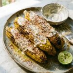 Grilled corn on the cob with charred kernels, butter, Parmesan, chili powder, and fresh lime wedges on a rustic wooden board