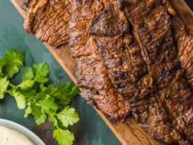 Grilled flank steak sliced thin and arranged on a wooden board surrounded by warm corn tortillas, fresh lime wedges, diced white onion, cilantro, and sliced avocado, shot on a rustic table with warm natural light