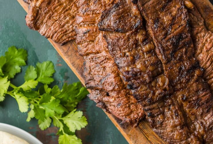 Grilled flank steak sliced thin and arranged on a wooden board surrounded by warm corn tortillas, fresh lime wedges, diced white onion, cilantro, and sliced avocado, shot on a rustic table with warm natural light