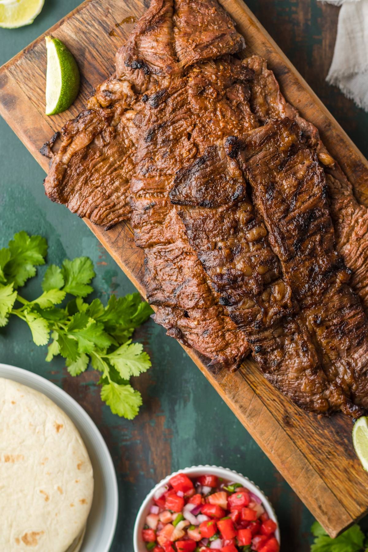 Grilled flank steak sliced thin and arranged on a wooden board surrounded by warm corn tortillas, fresh lime wedges, diced white onion, cilantro, and sliced avocado, shot on a rustic table with warm natural light
