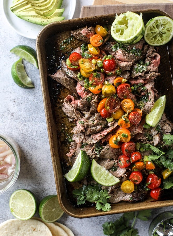 Overhead flat-lay of grilled flank steak marinade ingredients including lime halves, orange, garlic cloves, olive oil, smoked paprika, cumin, chili powder, soy sauce, and a raw flank steak on a white marble surface