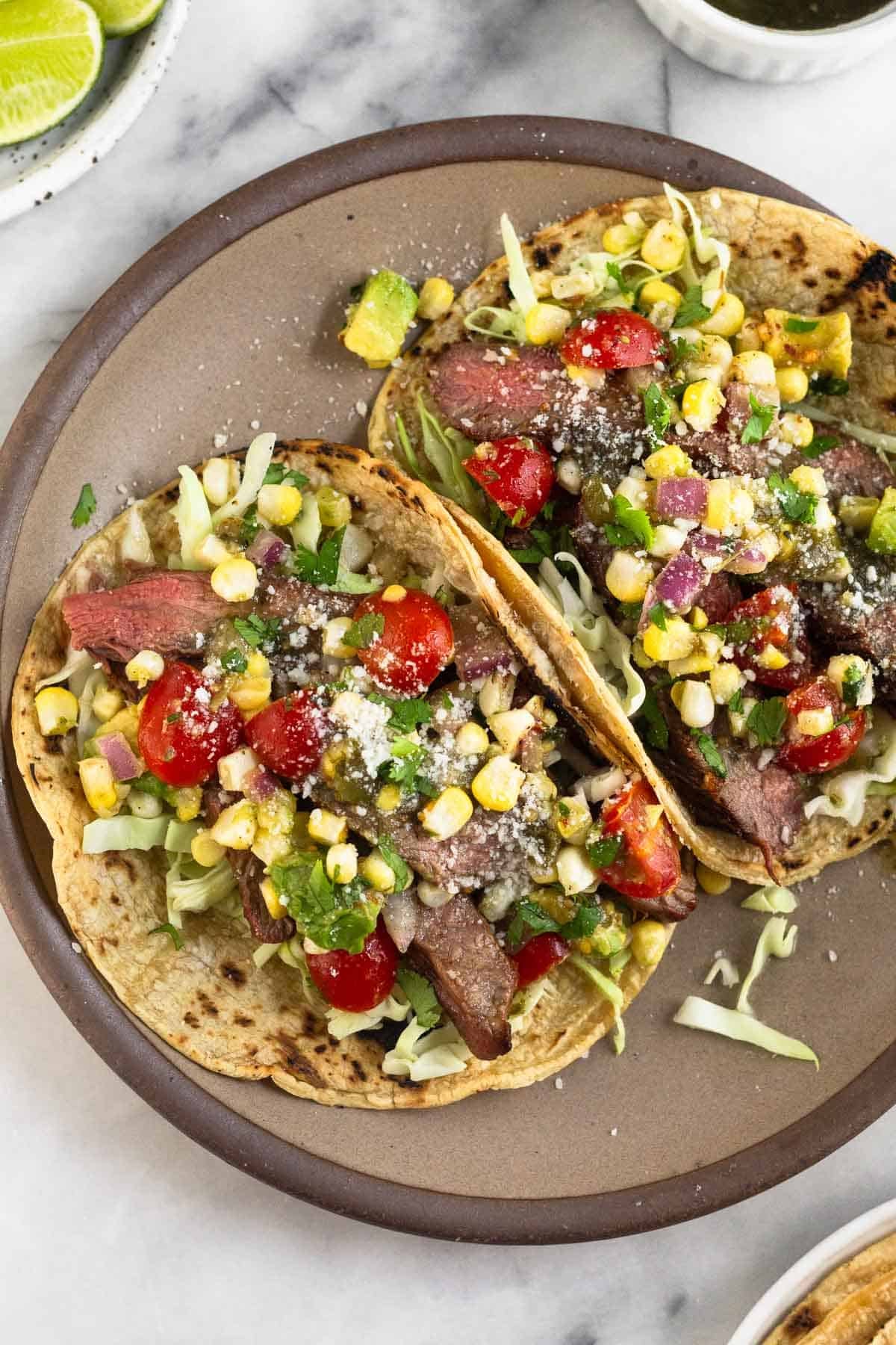 Close-up of grilled flank steak tacos assembled on corn tortillas with sliced steak, diced white onion, fresh cilantro, crumbled cotija cheese, sliced avocado, and a lime wedge on a vibrant ceramic plate