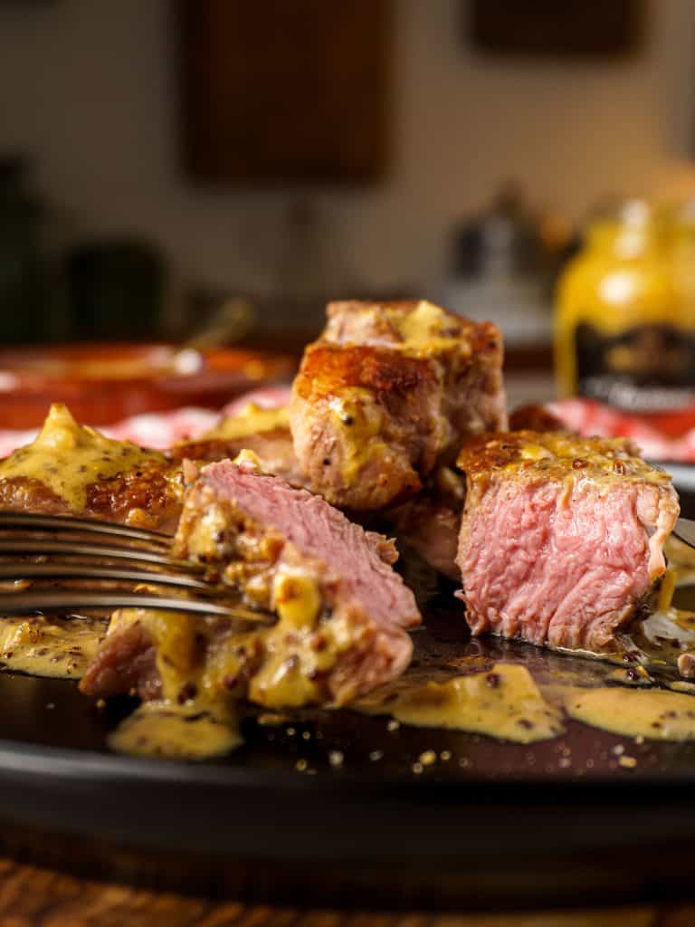 Close-up of sliced grilled pork tenderloin medallions fanned out on a white ceramic plate, showing juicy pink interior with golden-brown grill-marked exterior, garnished with fresh rosemary sprigs and a drizzle of pan sauce