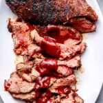 Perfectly grilled tri-tip steak sliced on a wooden cutting board with a smoky crust, pink interior, and fresh herbs scattered around