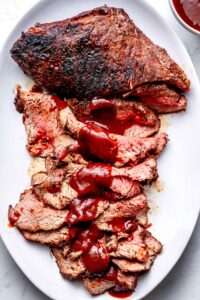 Perfectly grilled tri-tip steak sliced on a wooden cutting board with a smoky crust, pink interior, and fresh herbs scattered around