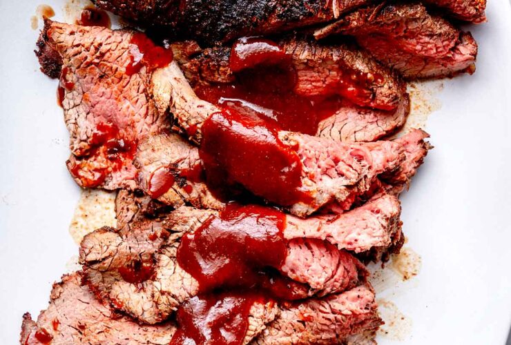Perfectly grilled tri-tip steak sliced on a wooden cutting board with a smoky crust, pink interior, and fresh herbs scattered around