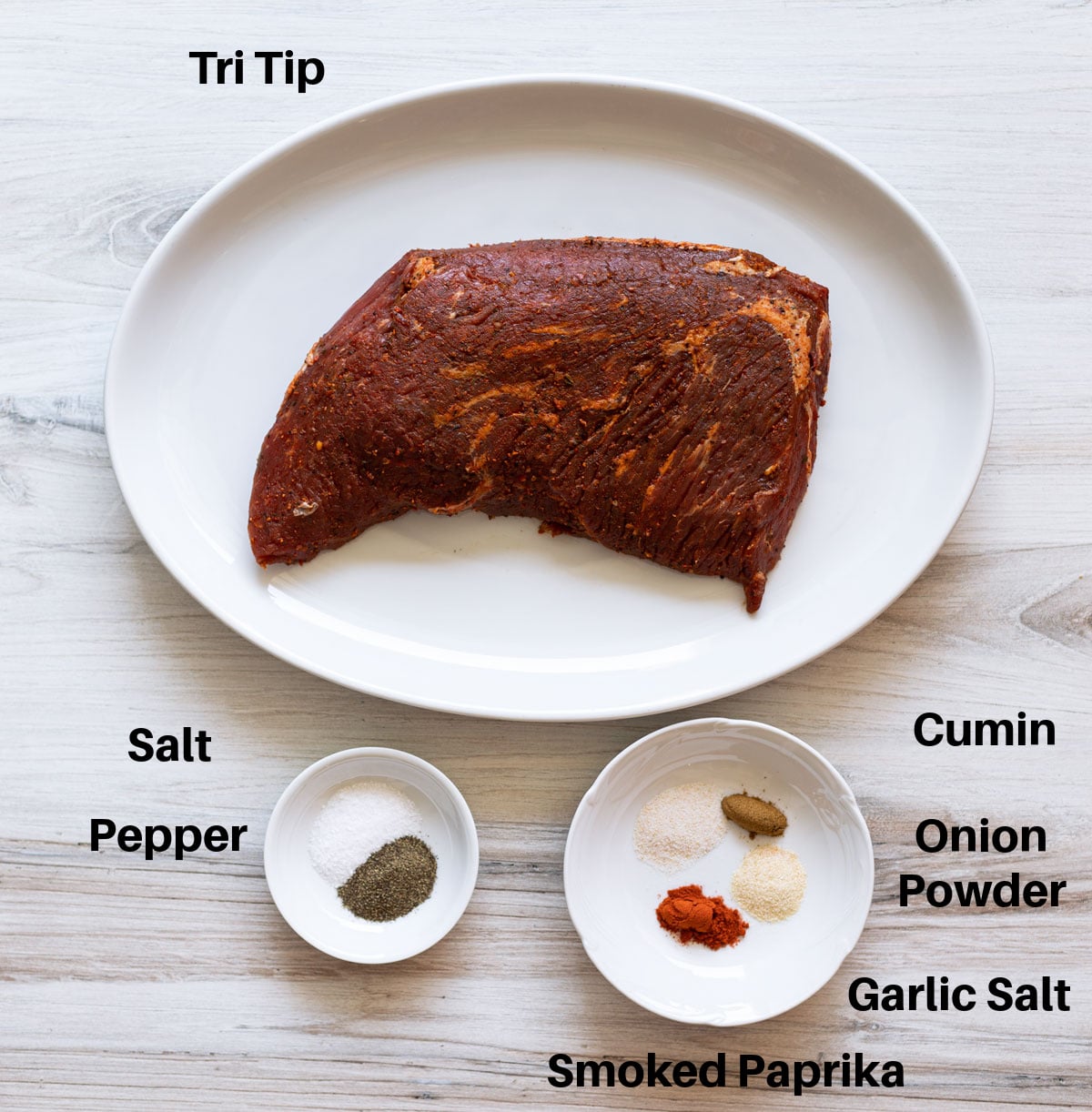 Tri-tip roast on a cutting board with dry rub ingredients including smoked paprika, garlic powder, salt, and olive oil arranged nearby